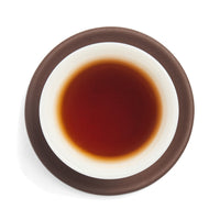 Brewed deep red sheng puerh tea liquor in a white porcelain cup on a clay coaster, seen from above