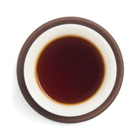 Brewed dark black tea (heicha) liquor in a white porcelain cup on a clay coaster, seen from above