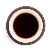 Brewed near-black black tea (heicha) liquor in a white porcelain cup on a clay coaster, seen from above